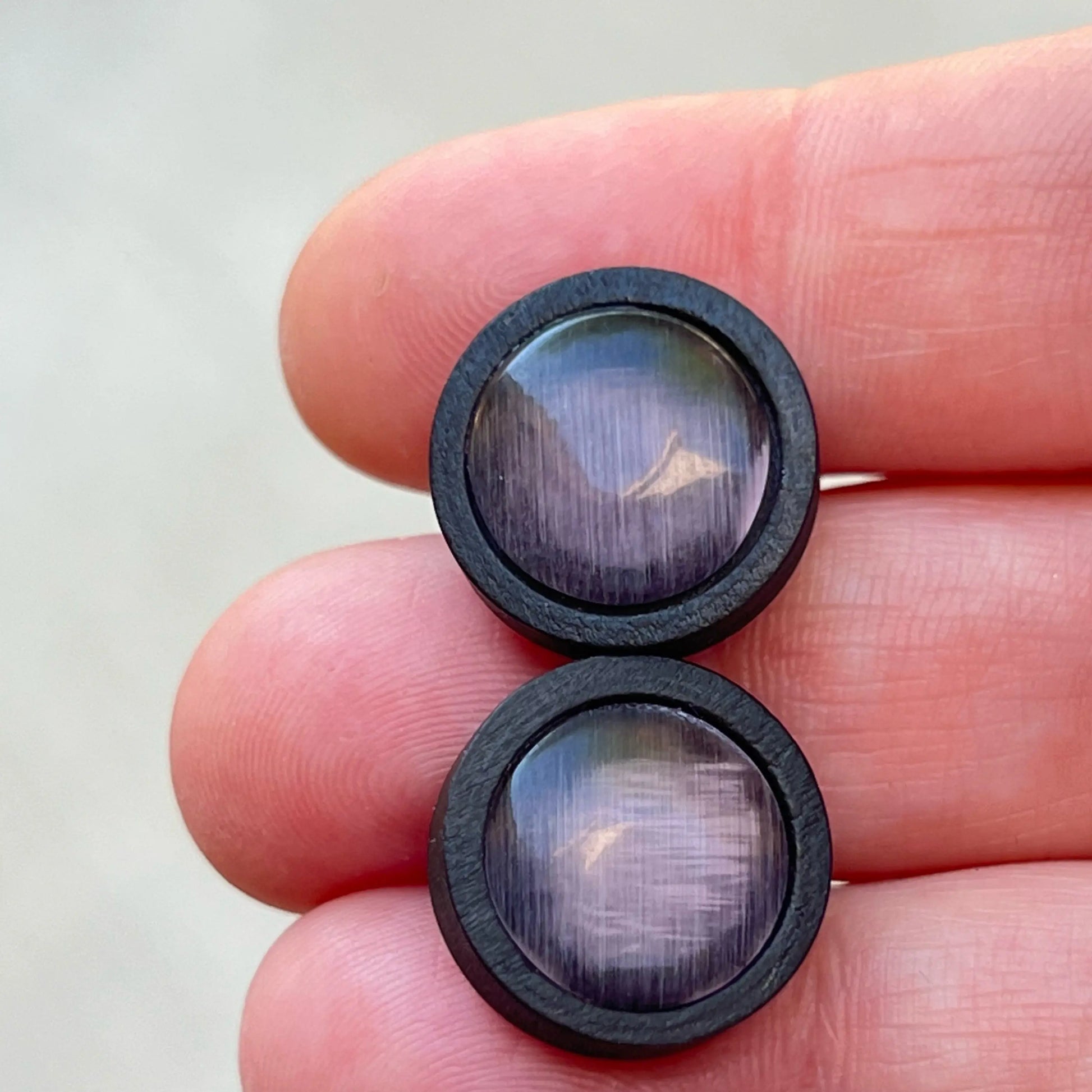 A pair of Purple Gray earrings with black wood rims held between fingers against a neutral background