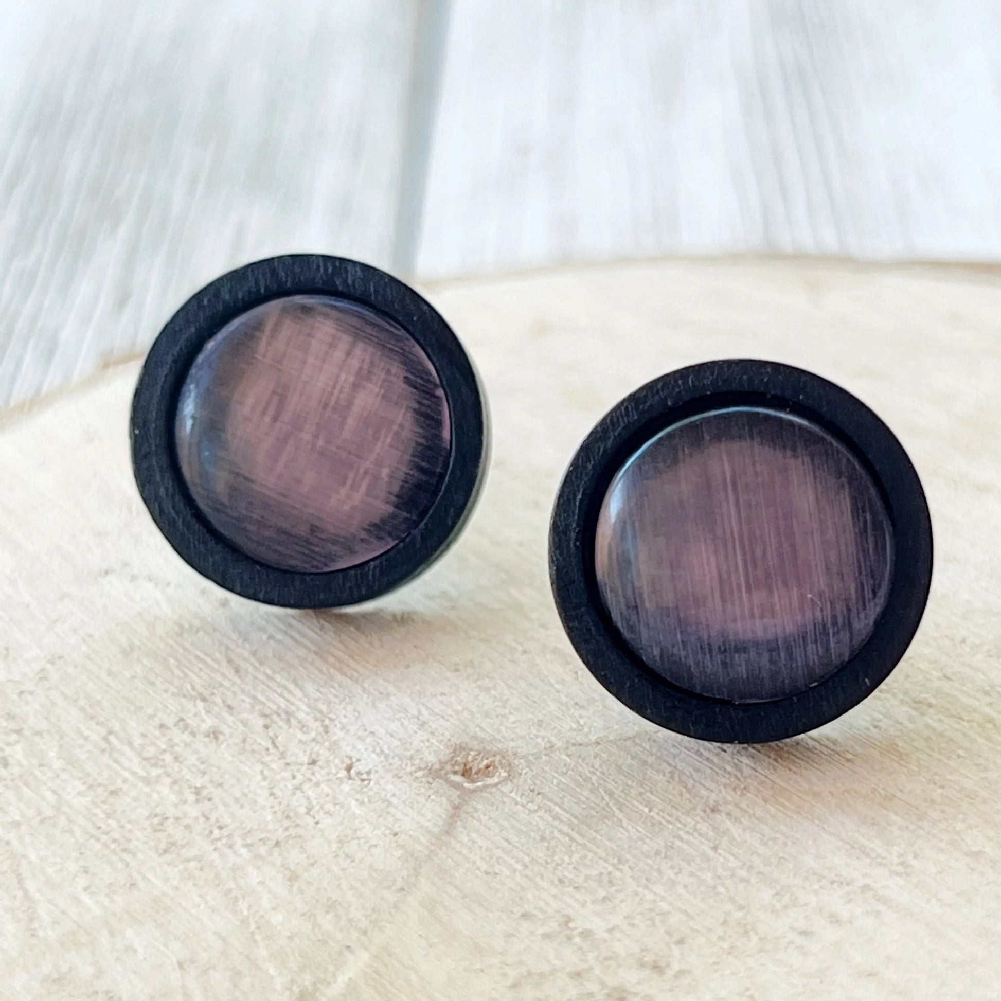 Round mauve gray glass stud earrings with black wood border displayed on light wood