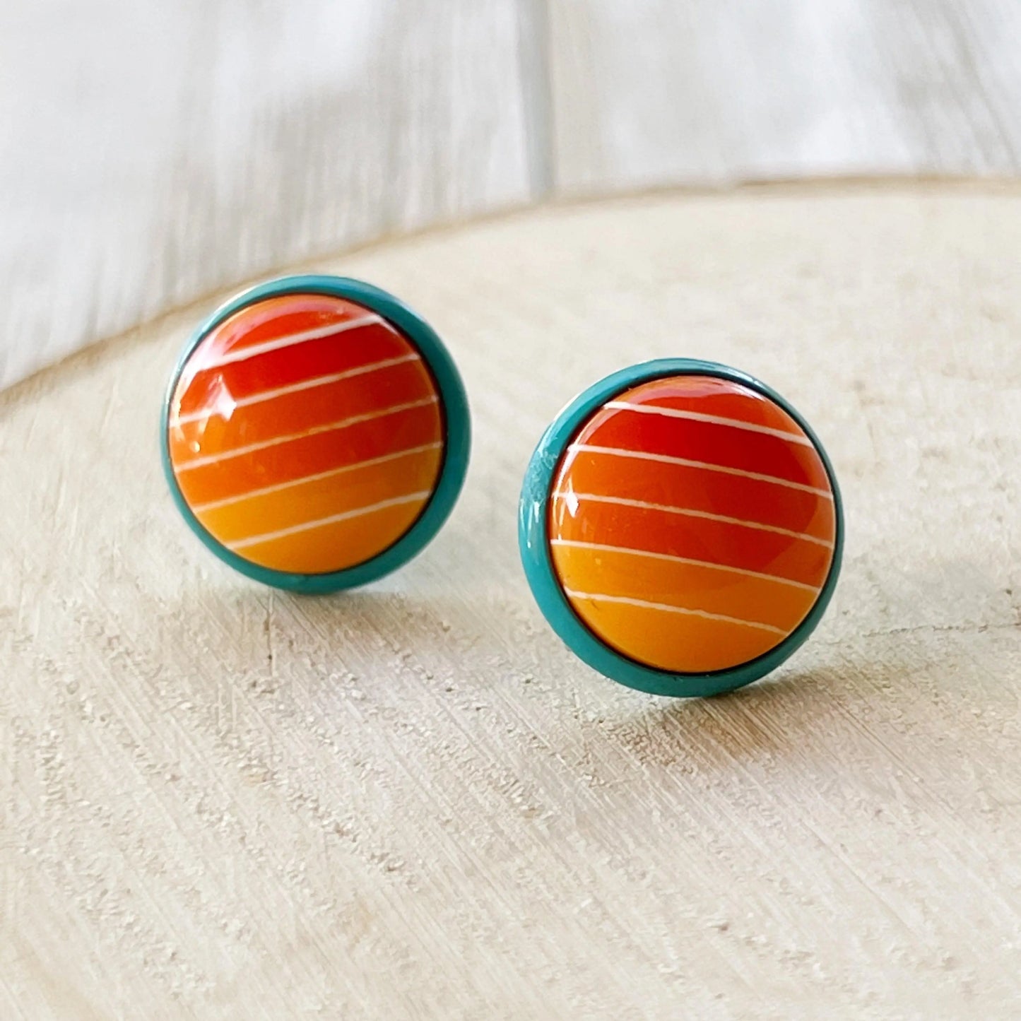 Round orange gradient striped stud earrings with teal borders displayed close up on a light wood background