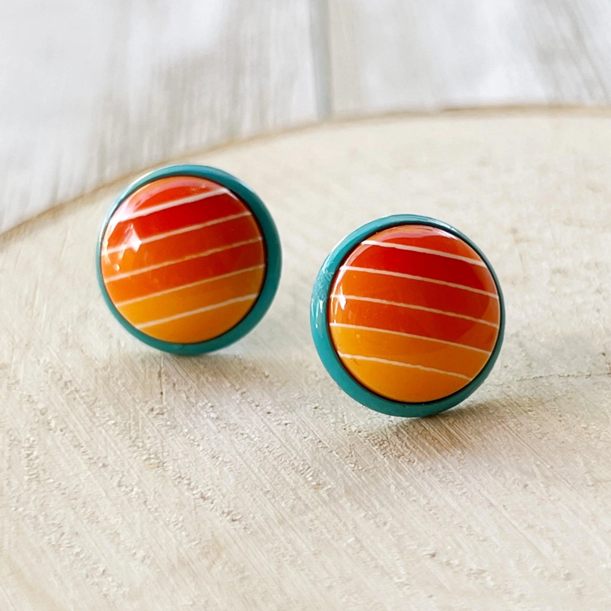 Round orange gradient striped stud earrings with teal borders displayed close up on a light wood background