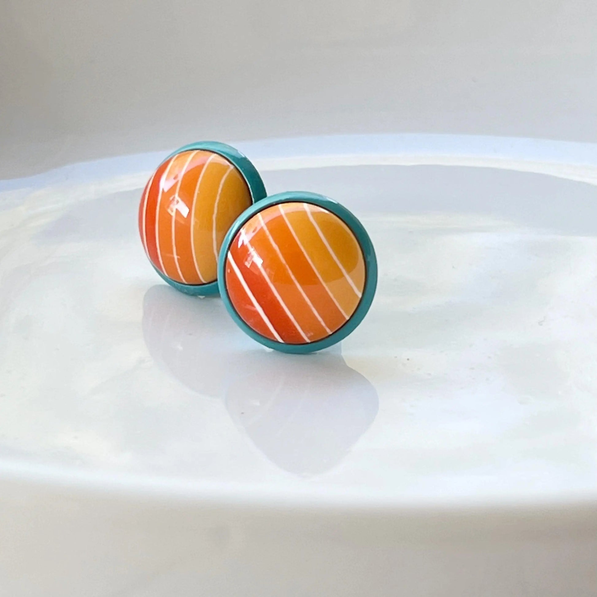 Round orange gradient striped stud earrings with teal borders displayed on a white dish