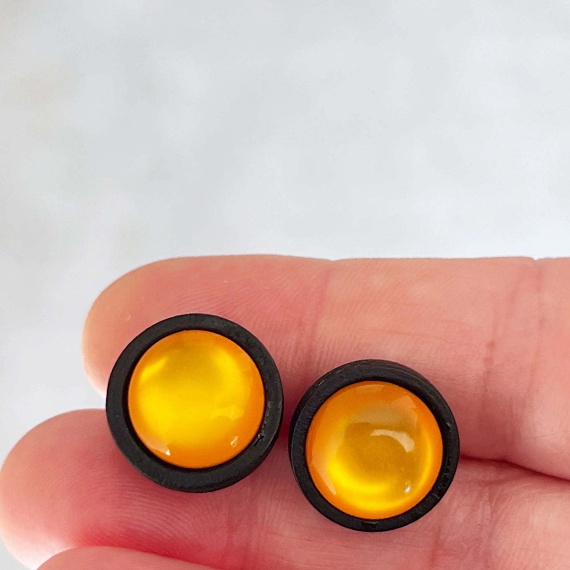 Round orange resin stud earrings with black wood border held on fingertips against a light background
