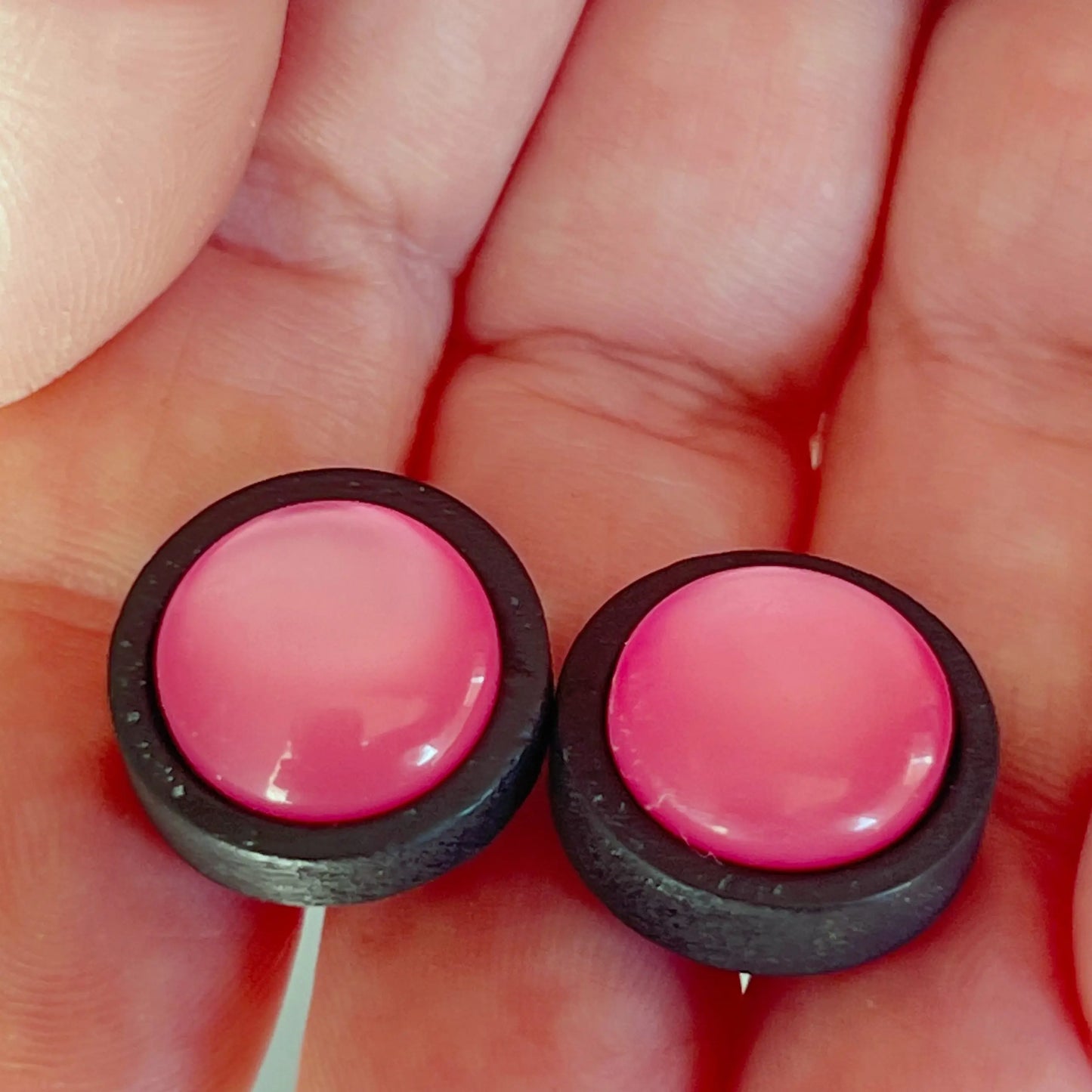 Two pink round earrings with black borders held between fingers