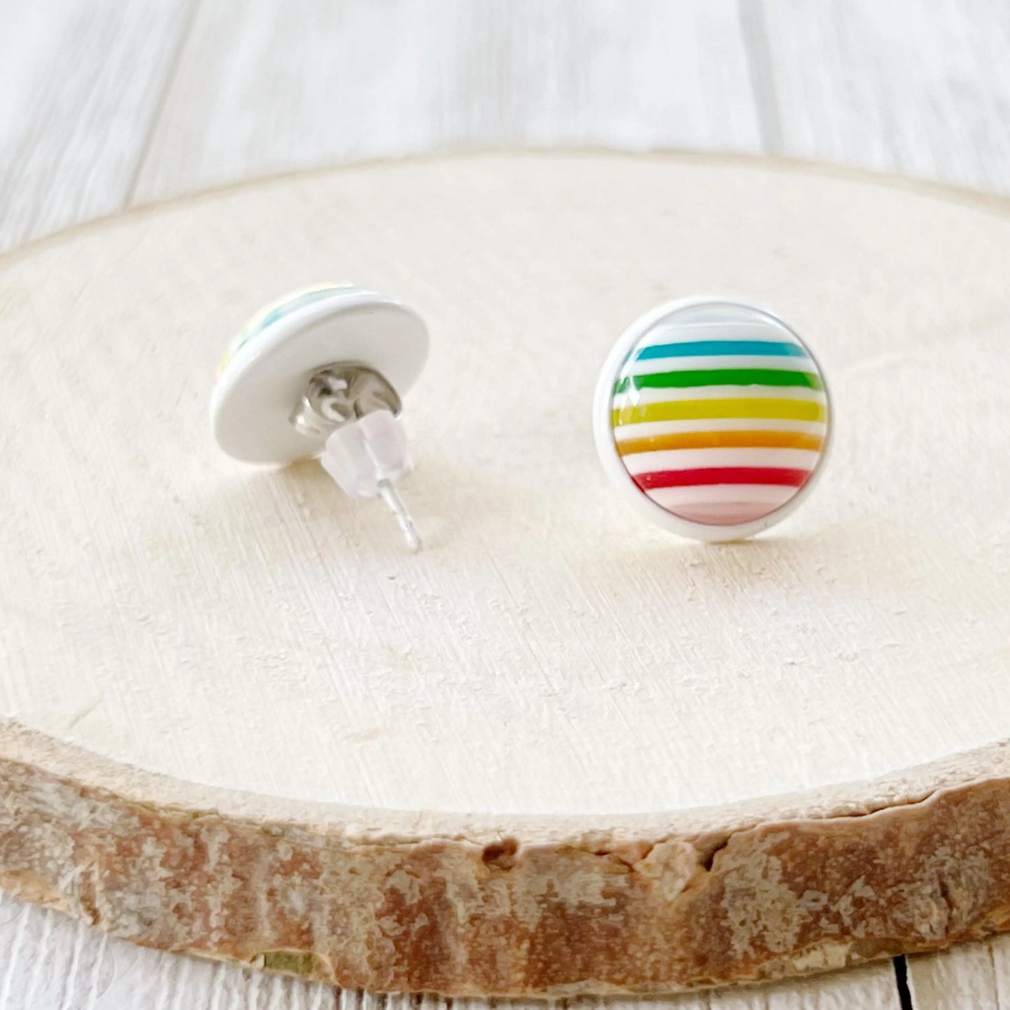 Round rainbow stripe stud earrings in a white setting displayed on a wood slice