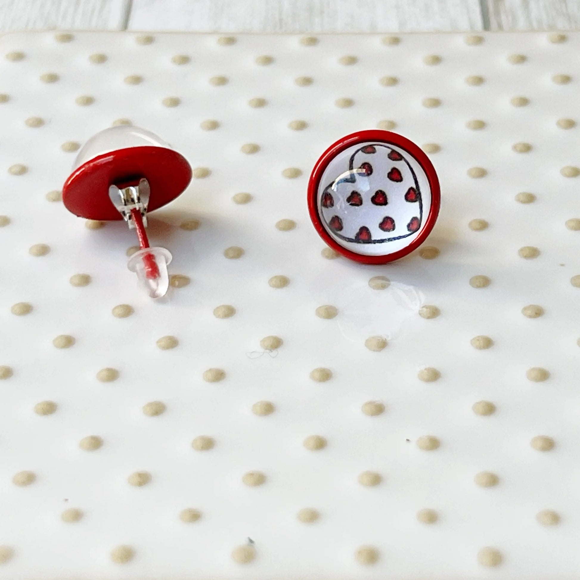 Back of round red stud earrings with white heart artwork and tiny red heart details displayed on a glass tray