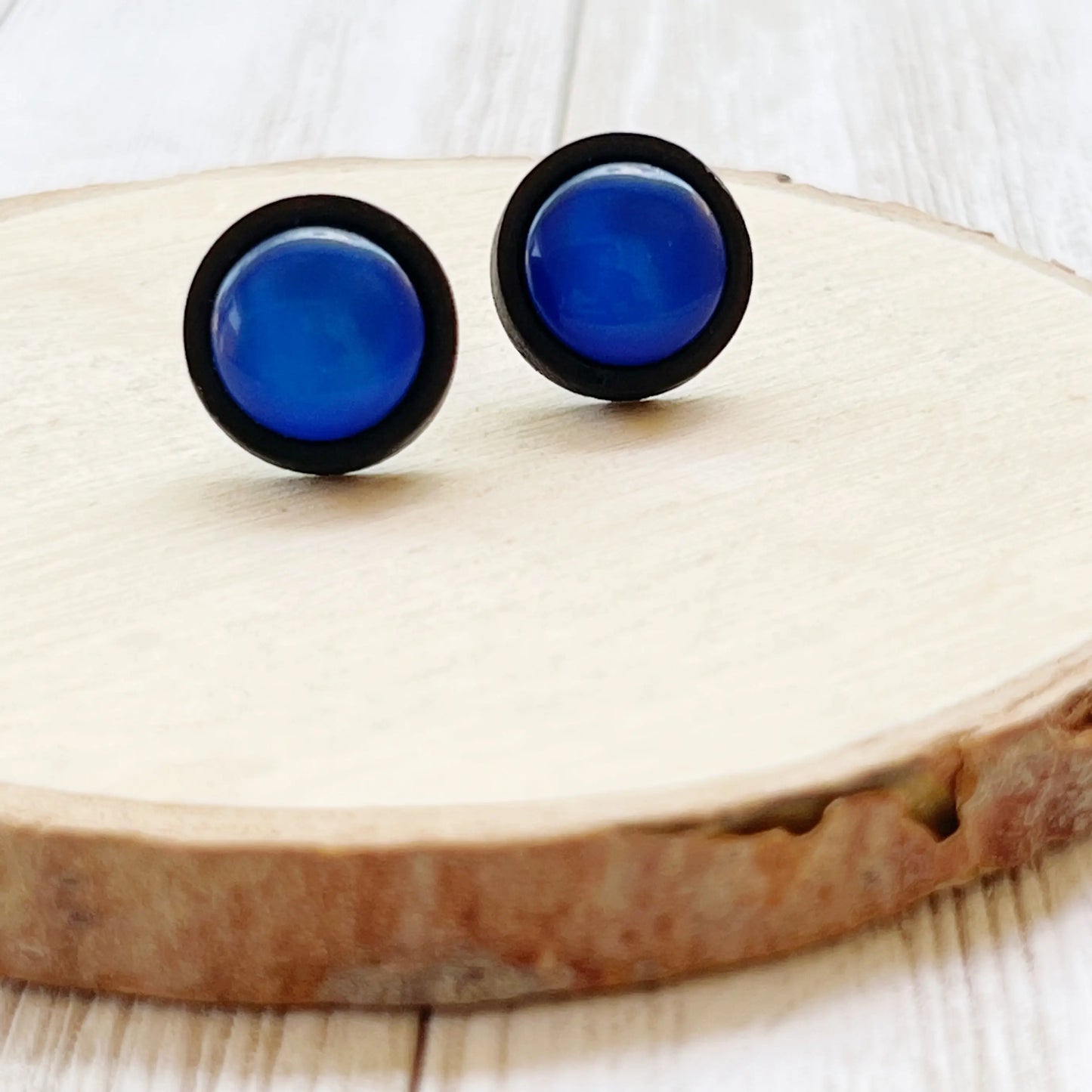 Round royal blue resin stud earrings with black wood borders displayed on a light wood slice