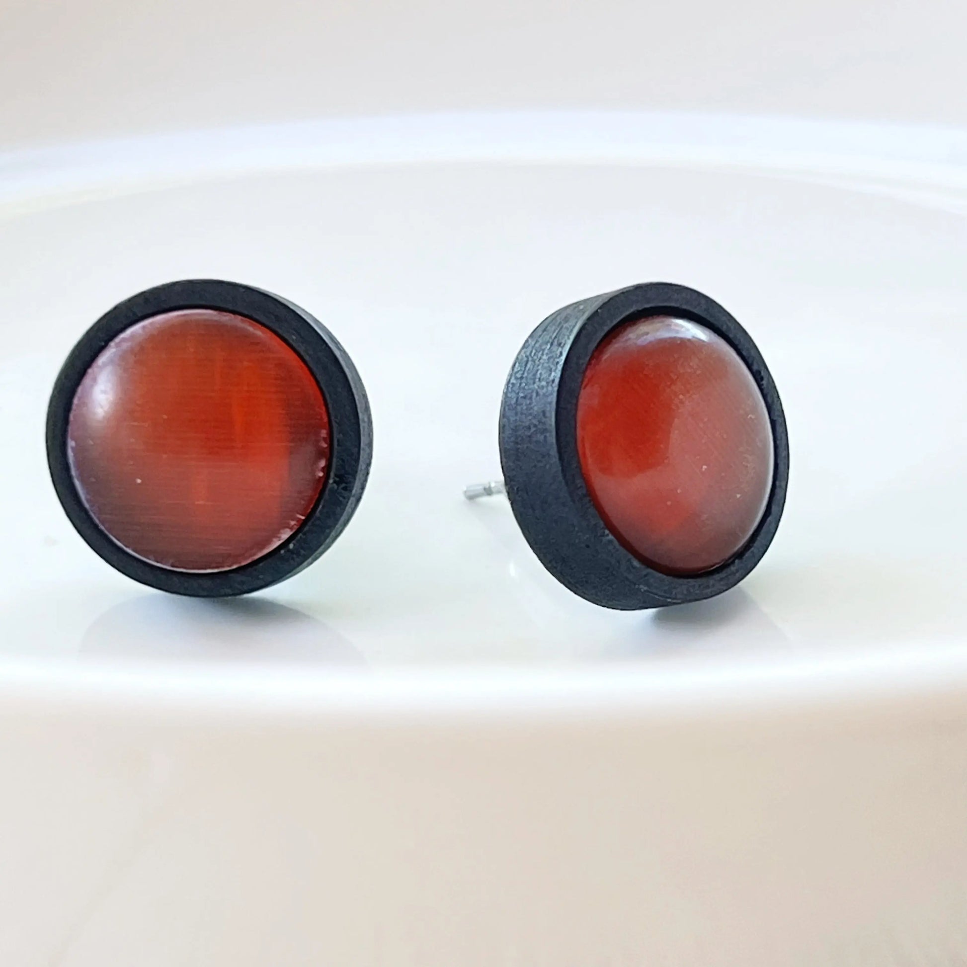 Round rust orange glass stud earrings with black wood border displayed on a white dish