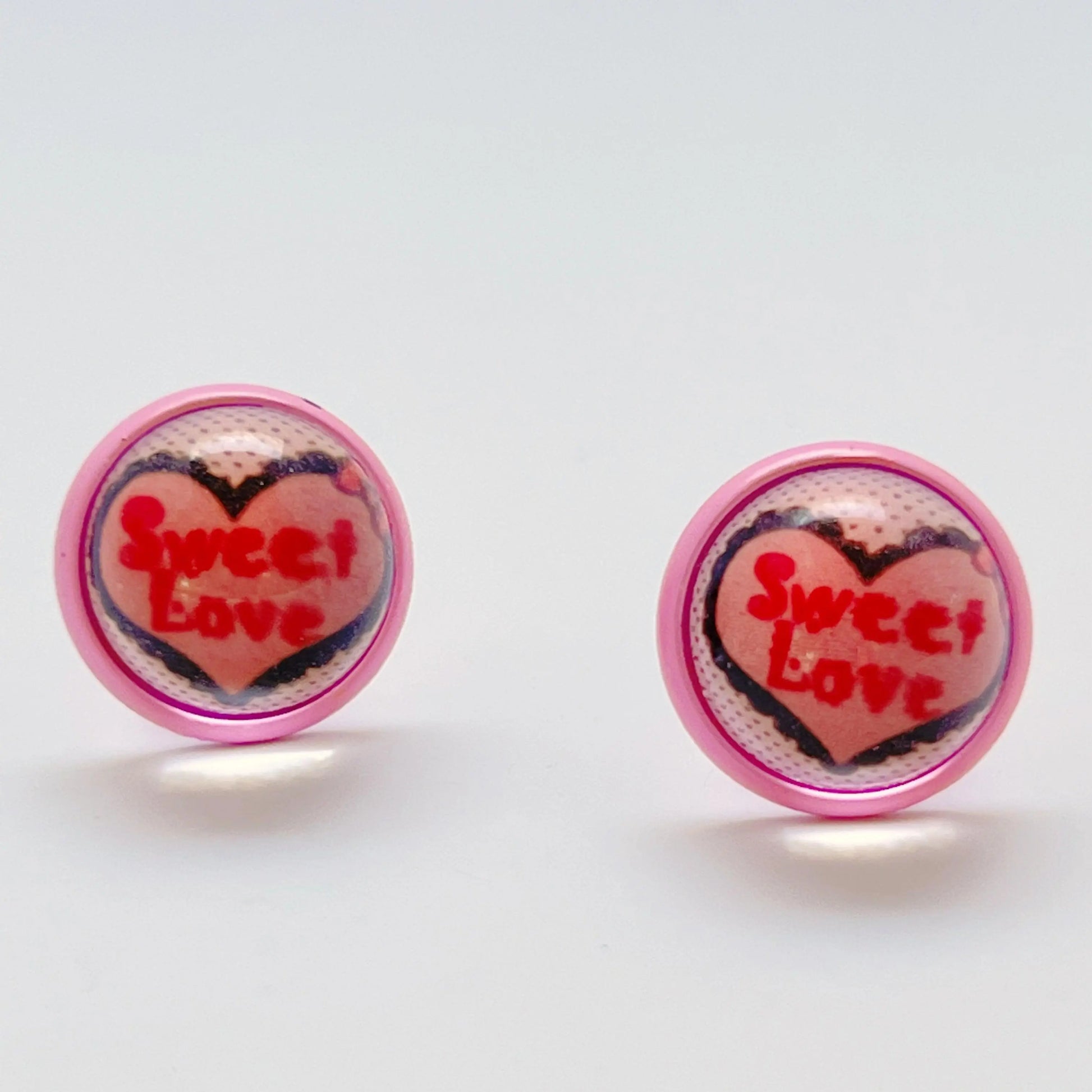 Round pink stud earrings with Sweet Love conversation heart artwork on a white background