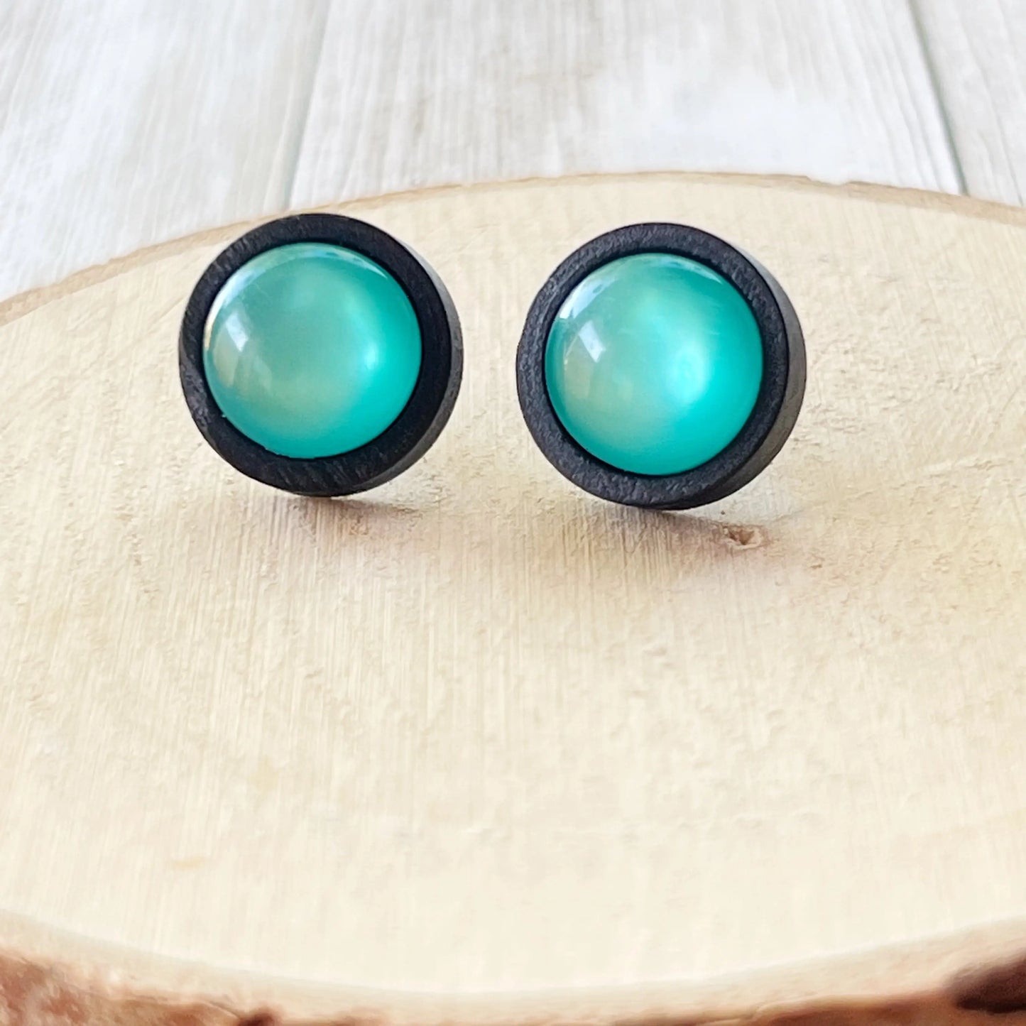 Round black wood stud earrings with glossy teal resin centers shown on wood slice