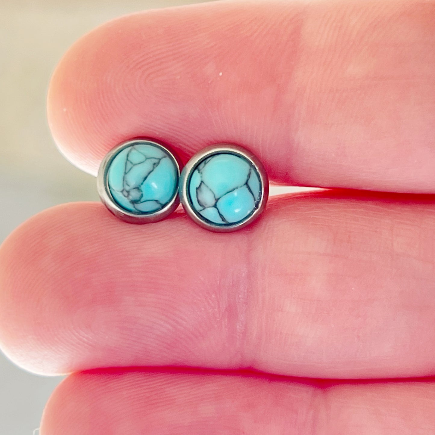 Small round turquoise stud earrings in a silver-tone setting shown close up on fingertips